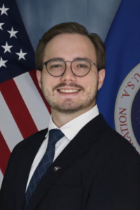 Alexander Bahrim smiling in a dark suit and tie, wearing glasses, with the U.S. and NASA flags in the background.